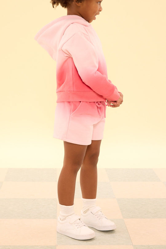 Child wearing a pink and purple gradient hoodie and shorts on a light background