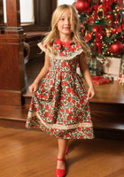 A young girl wearing the Ivy City Co Mini Aster Dress in Red Roses and matching red shoes stands smiling in front of a decorated Christmas tree with presents underneath.