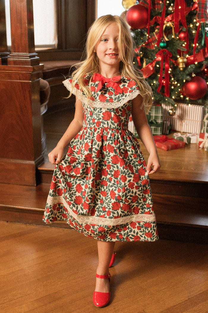 A young girl wearing the Ivy City Co Mini Aster Dress in Red Roses and matching red shoes stands smiling in front of a decorated Christmas tree with presents underneath.