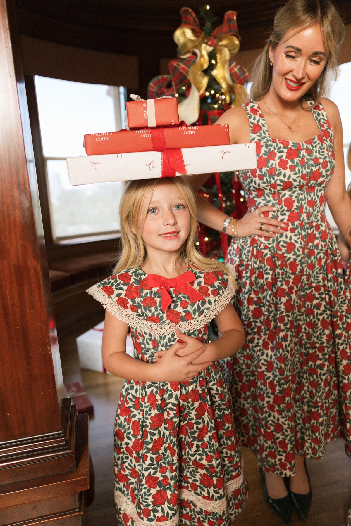 A smiling girl balances three wrapped gifts on her head beside a woman; both are wearing the Ivy City Co Mini Aster Dress in Red Roses, featuring delicate lace trim and a festive floral design. A decorated Christmas tree stands in the background.