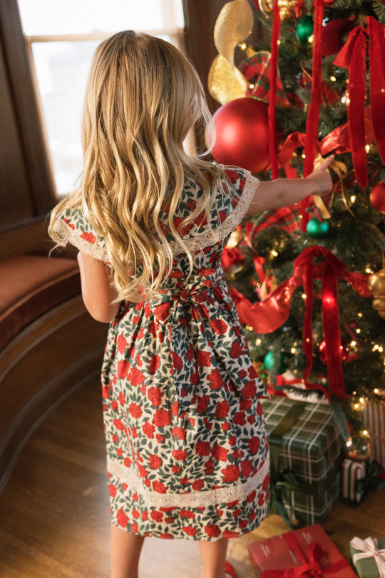 A young girl wearing the Ivy City Co Mini Aster Dress in Red Roses, featuring delicate lace trim, stands beside a decorated Christmas tree and reaches up to hang an ornament, with presents stacked underneath the tree.