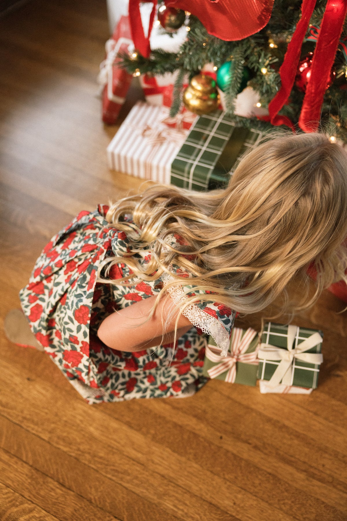 A young girl wearing the Ivy City Co Mini Aster Dress in Red Roses, featuring delicate lace trim, kneels on a wooden floor as she reaches for wrapped gifts beneath a decorated Christmas tree.
