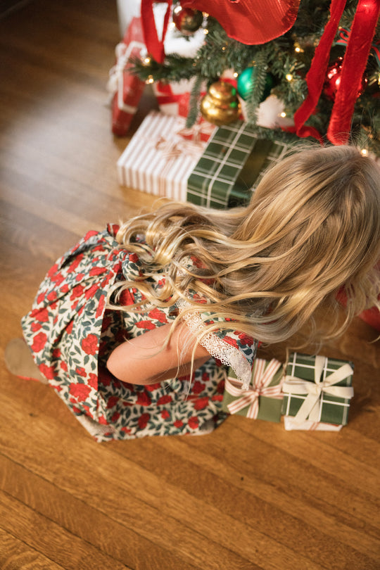 A young girl wearing the Ivy City Co Mini Aster Dress in Red Roses, featuring delicate lace trim, kneels on a wooden floor as she reaches for wrapped gifts beneath a decorated Christmas tree.