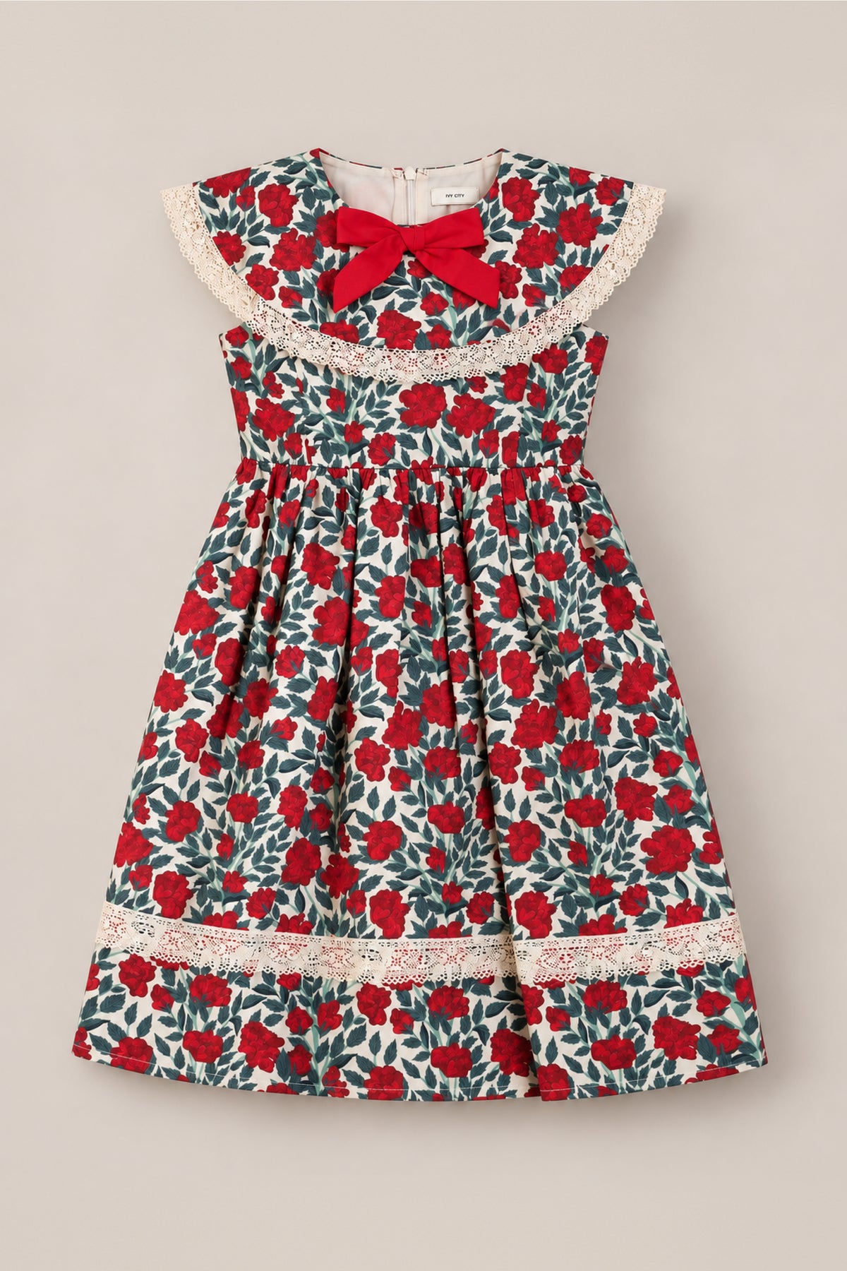 The Ivy City Co Mini Aster Dress in Red Roses features a sleeveless design with a vibrant red and green floral pattern, delicate lace trim, a large round lace collar, and a charming red bow at the neckline. Displayed flat against a neutral background, this lovely holiday dress is perfect for any special occasion.