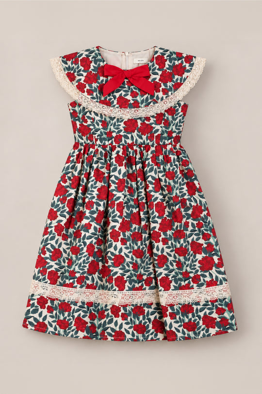The Ivy City Co Mini Aster Dress in Red Roses features a sleeveless design with a vibrant red and green floral pattern, delicate lace trim, a large round lace collar, and a charming red bow at the neckline. Displayed flat against a neutral background, this lovely holiday dress is perfect for any special occasion.