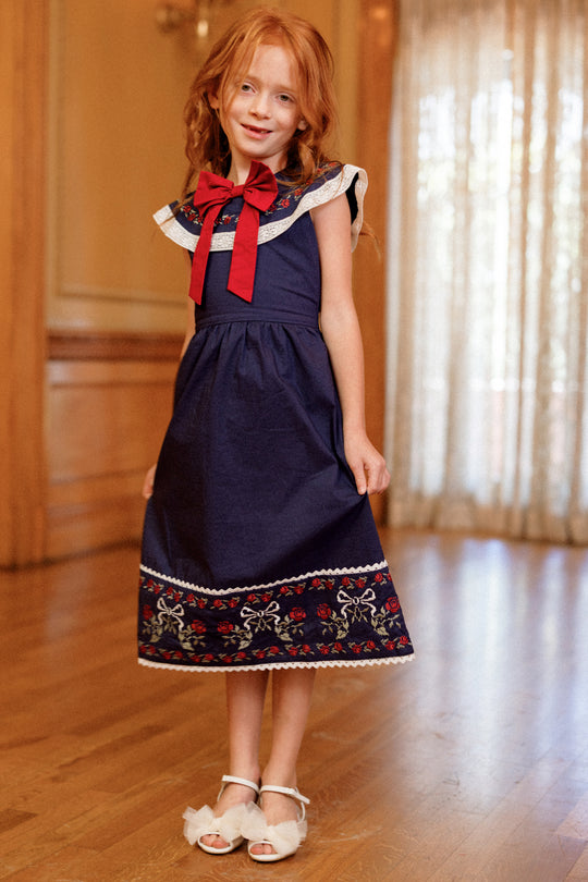 A young girl with red hair stands smiling on a wooden floor in a warmly lit room with sheer curtains, wearing the Ivy City Co Mini Aster Dress in Sapphire Rosette Embroidery, which features delicate floral and rosette detailing, topped with a large red bow and paired with white sandals.