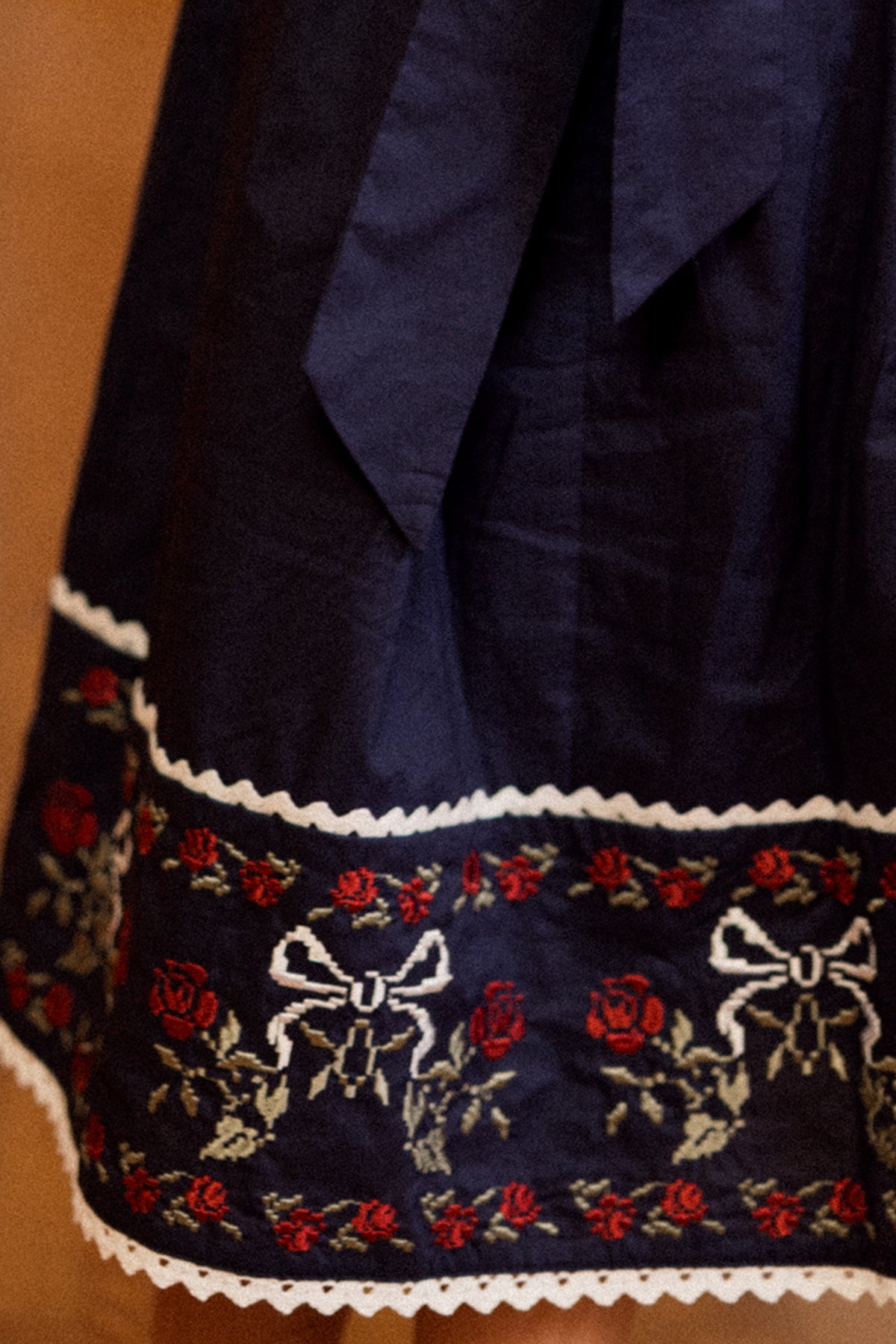 A close-up of the Mini Aster Dress in Sapphire Rosette Embroidery by Ivy City Co showcases its dark blue fabric with a border of red roses, green leaves, white bows, and delicate white lace trim. The gathered fabric has a subtle sheen, making it perfect for holiday portraits or coordinating family dress sets.