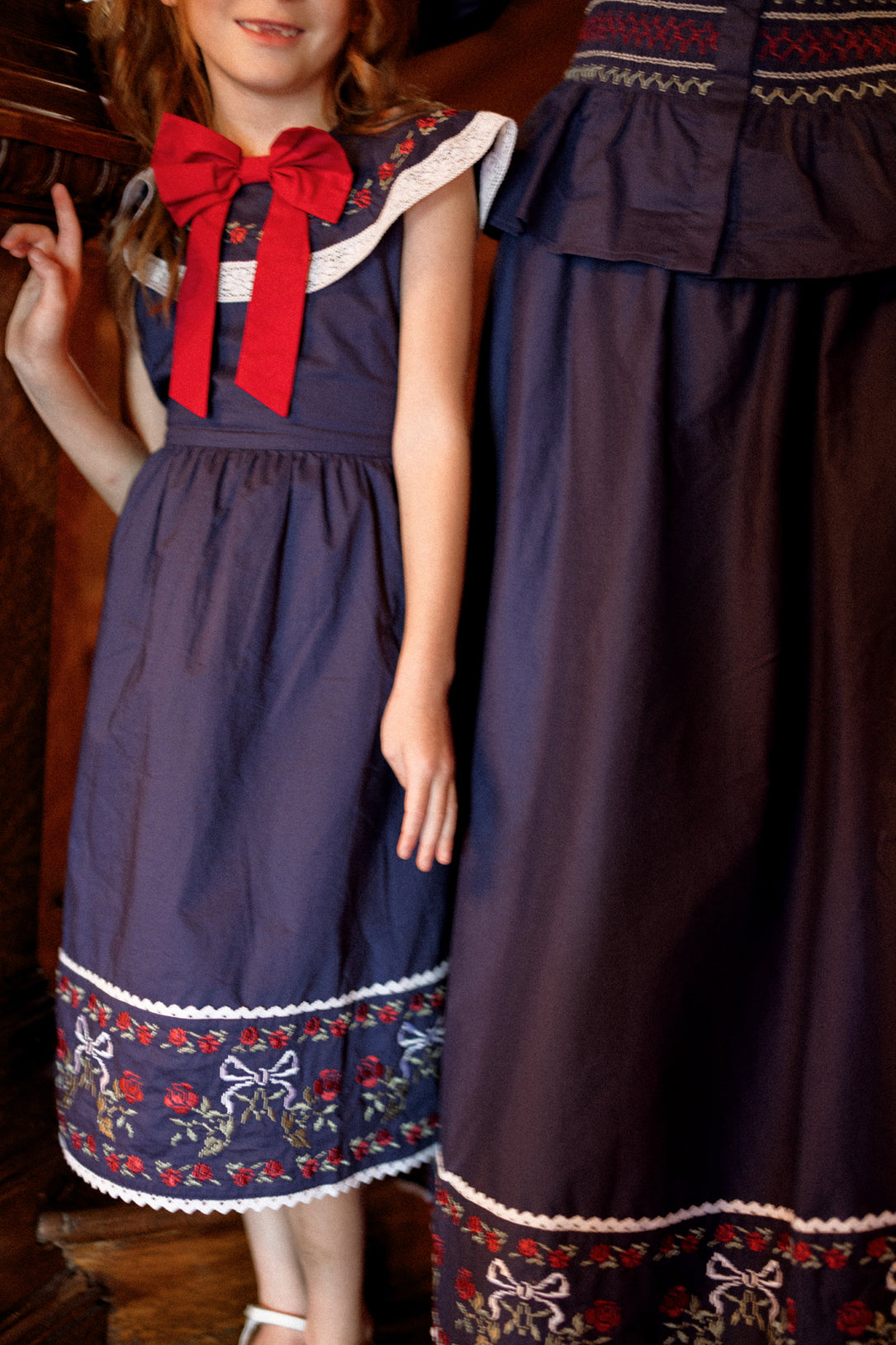 A young girl and an adult, both wearing matching Ivy City Co Mini Aster Dresses in Sapphire Rosette Embroidery, stand side by side indoors. Only the girl’s face is partially visible; both are cropped at the shoulders.