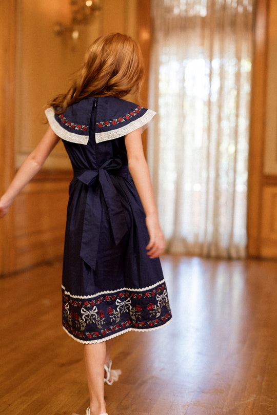 A young girl with long red hair, wearing the Ivy City Co Mini Aster Dress in Sapphire Rosette Embroidery, walks barefoot across a wooden floor toward a sheer curtain.