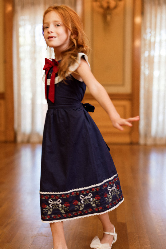A young girl with red hair twirls on a wooden floor in a softly lit room with large windows and light curtains, wearing the Ivy City Co Mini Aster Dress in Sapphire Rosette Embroidery.