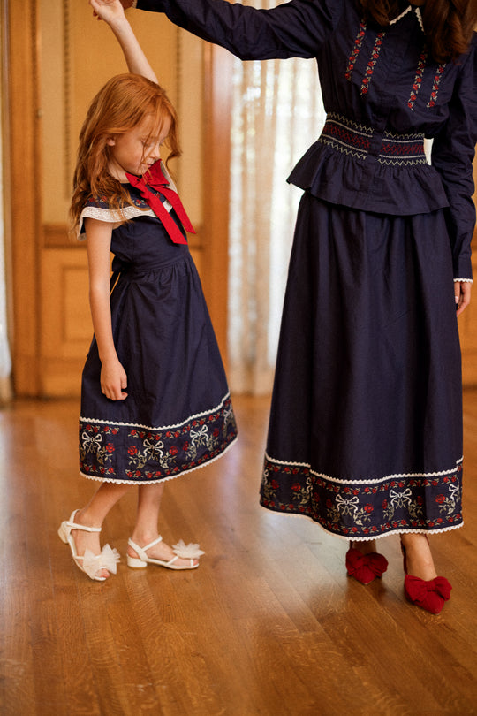 A red-haired girl and a woman stand together indoors, both wearing Ivy City Co’s Mini Aster Dress in Sapphire Rosette Embroidery. The matching family dresses feature navy blue fabric, white lace trim, vibrant red floral embroidery, and delicate rosette embroidery accents along the hems. The girl pairs her dress with white sandals, while the woman complements her look with red heels adorned with bows—making these outfits perfect for a memorable holiday portrait.