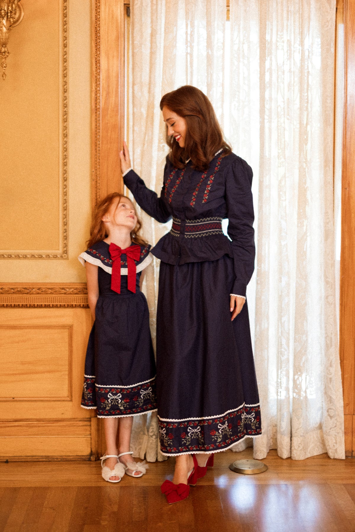A woman and a girl beam together by a window trimmed in wood and framed with lace curtains, both dressed in Ivy City Co’s Mini Aster Dress in Sapphire Rosette Embroidery—making the perfect holiday portrait in their matching family outfits.