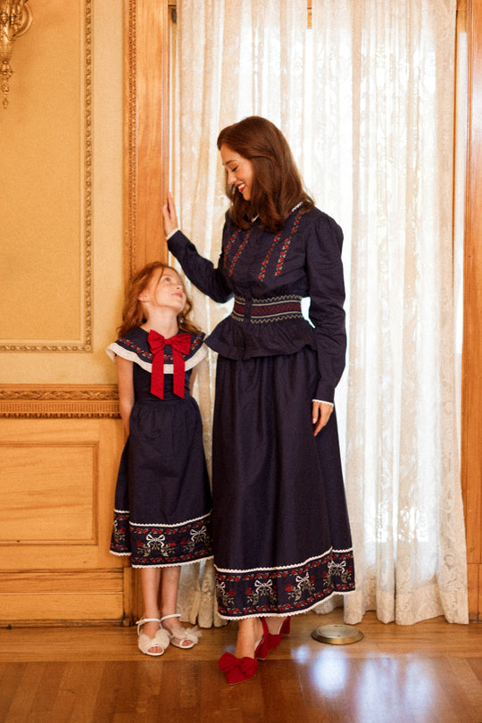 A woman and a girl beam together by a window trimmed in wood and framed with lace curtains, both dressed in Ivy City Co’s Mini Aster Dress in Sapphire Rosette Embroidery—making the perfect holiday portrait in their matching family outfits.