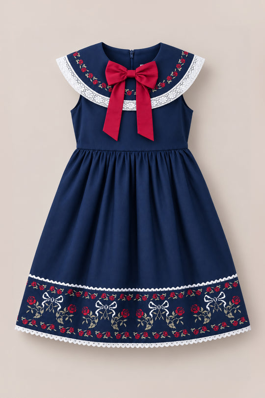 The Mini Aster Dress in Sapphire Rosette Embroidery by Ivy City Co features a navy blue sleeveless design with a wide collar, red bow, white lace trim, and delicate rosette embroidery along the collar and hem—perfect for holiday portraits or coordinating family dress looks.