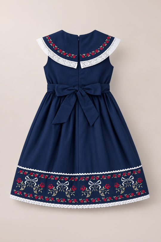 The Mini Aster Dress in Sapphire Rosette Embroidery by Ivy City Co features a navy blue sleeveless design with a white lace-trimmed collar, delicate floral and rosette embroidery, a sash tied in a bow at the waist, and embroidered flowers and bows along the hem—making it the perfect matching family dress for special occasions.