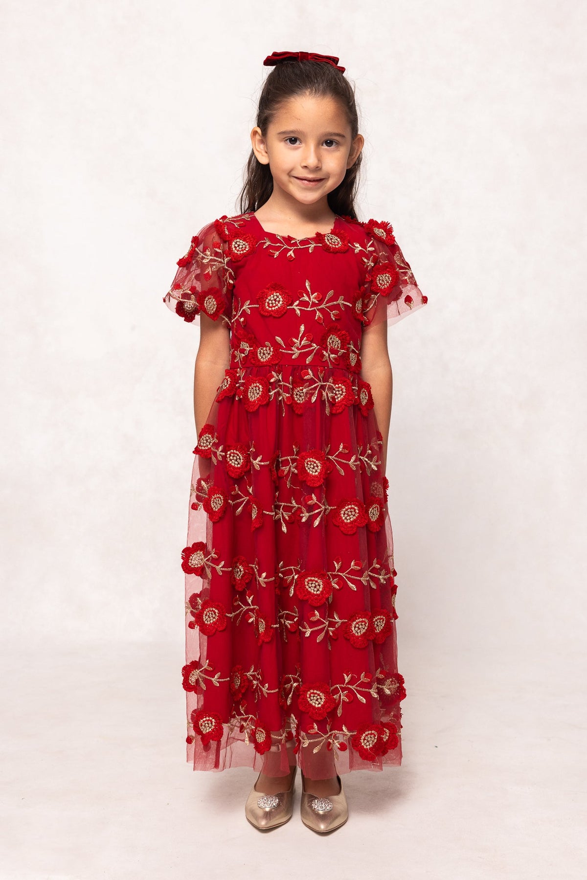 A young girl stands and smiles, dressed in the Ivy City Co Mini Athena Dress in Red—a beautifully embroidered short-sleeve maxi—paired with gold shoes and a coordinating red headband against a plain light background. Perfect as a holiday outfit or for matching in a mommy and me dress set.