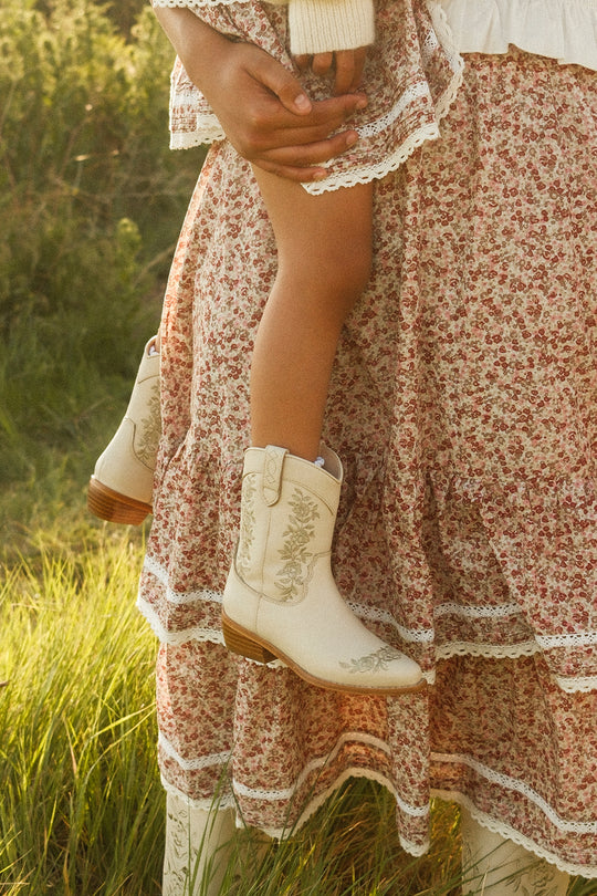 A person in a floral skirt holds a child wearing Ivy City Co's Mini Avery Boot in Cream, standing together outdoors on grass.