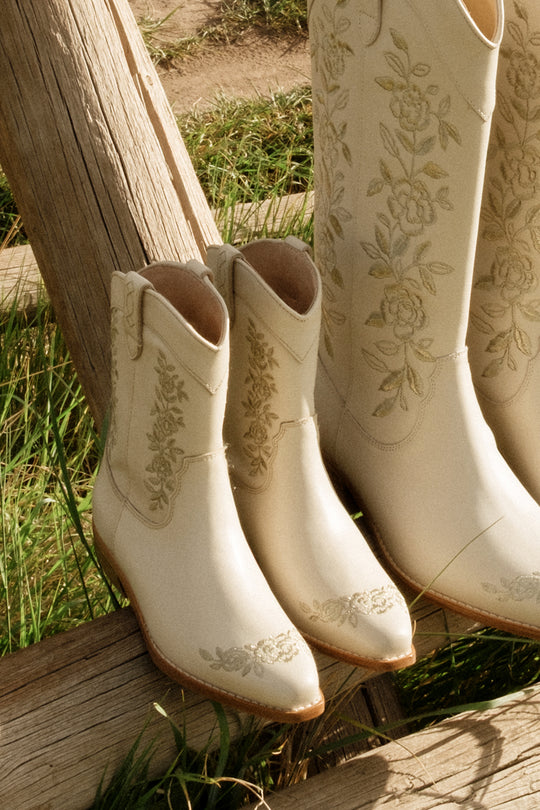 The Ivy City Co Mini Avery Boot in Cream for toddlers, featuring floral embroidery, sits next to matching taller cream boots on a wooden bench outdoors with green grass in the background.