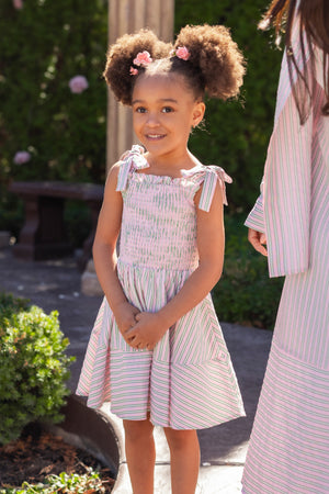A young girl with curly hair smiles outdoors in the Ivy City Co Mini Bluebell Dress in Green & Pink Pinstripe, with greenery behind her. An adult in a matching outfit stands beside her, partially out of frame.