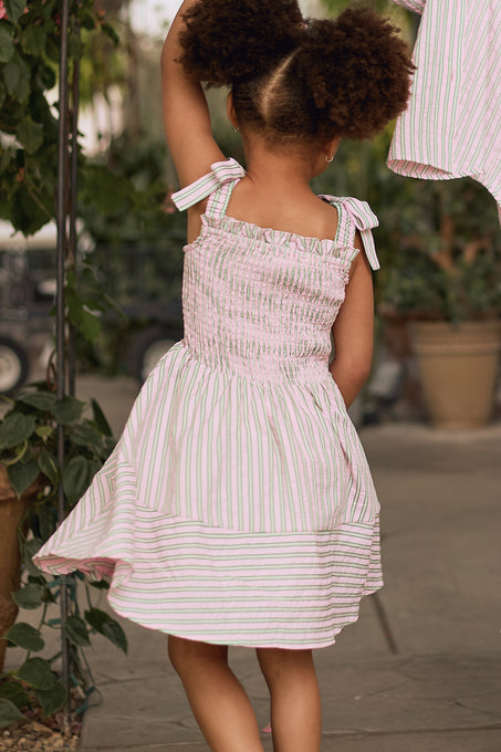 A young child with curly hair, seen from behind, spins playfully outdoors near potted plants while wearing the Mini Bluebell Dress in Green & Pink Pinstripe by Ivy City Co.