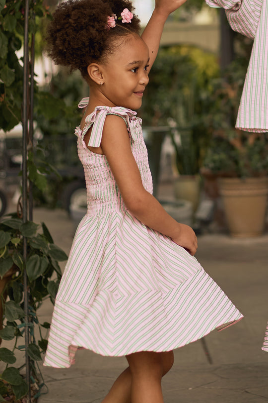 A smiling young girl with curly hair and pink flower clips twirls outdoors among potted plants, wearing Ivy City Co’s sleeveless Mini Bluebell Dress in Green & Pink Pinstripe, complete with tie straps.