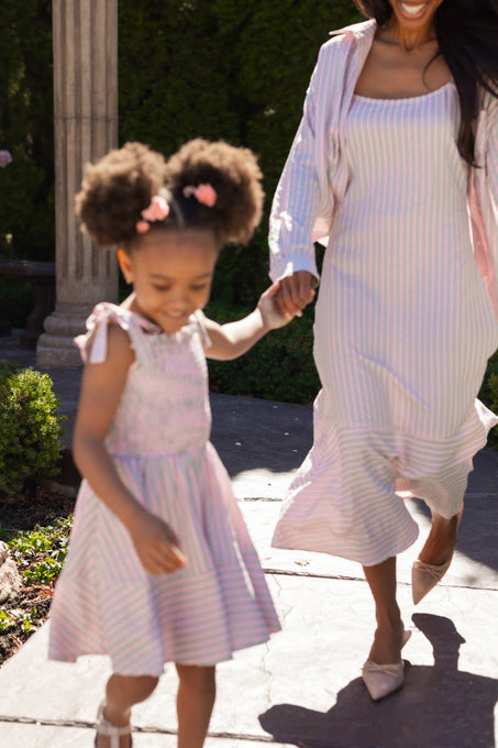 A woman and a young girl, both wearing Ivy City Co's Mini Bluebell Dress in Green & Pink Pinstripe, hold hands and smile as they walk outdoors on a sunny day; the girl’s puffy hair is styled with pink bows. Only part of the woman's face is visible.