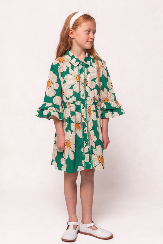 A young girl with long red hair wears the Ivy City Co Mini Bowie Dress in Green with White Floral, paired with white shoes and a matching headband, standing against a plain light background.