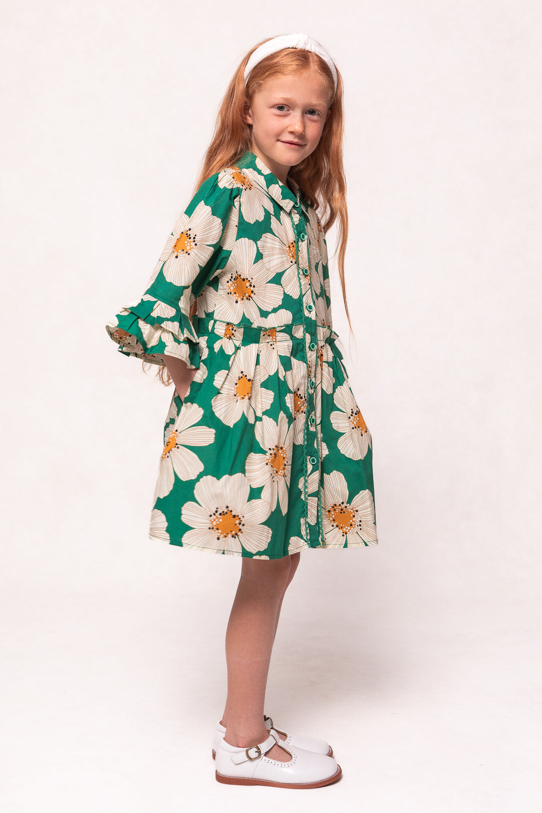 A young girl with long red hair wears the Ivy City Co Mini Bowie Dress in Green with White Floral, styled with a white headband and shoes, standing sideways and smiling against a plain white background.