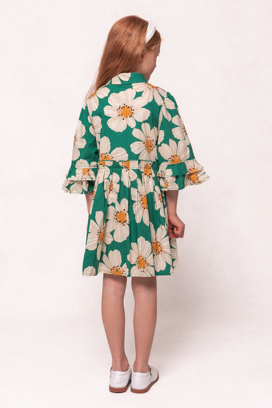 A young girl with long red hair, seen from behind, wears the Ivy City Co Mini Bowie Dress in Green with White Floral, paired with white shoes and a white headband.
