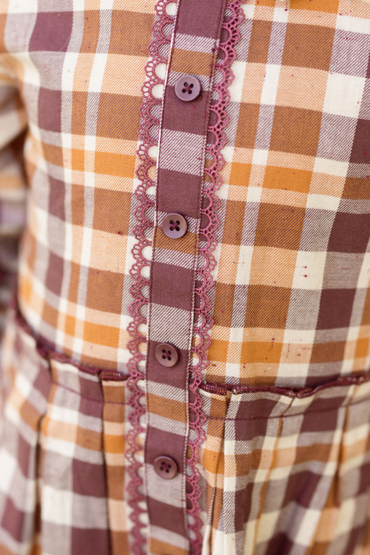 Close-up of the Mini Bowie Long Sleeve Dress in Spiced Plaid by Ivy City Co, a cotton girls’ dress in brown, orange, and white plaid with purple buttons and lace trim along the button placket.