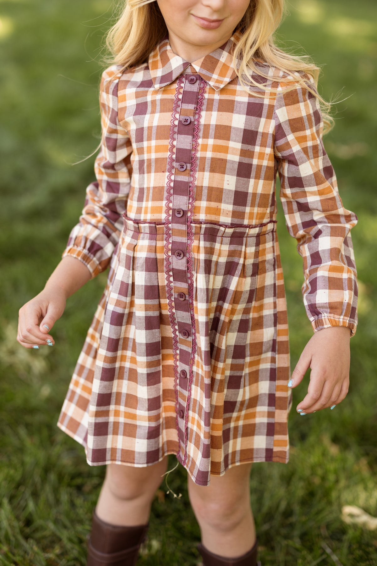 A young girl stands outside on green grass wearing the Ivy City Co Mini Bowie Long Sleeve Dress in Spiced Plaid, featuring a brown, orange, and white plaid pattern with buttons down the front.