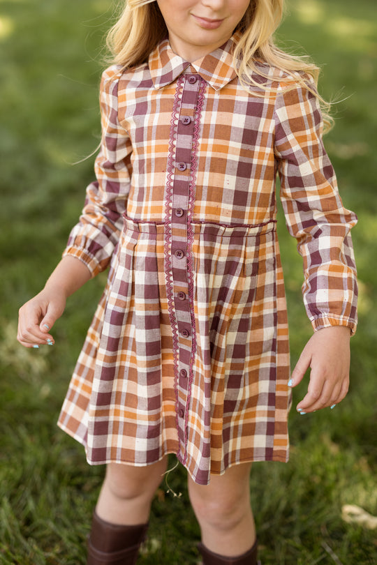 A young girl stands outside on green grass wearing the Ivy City Co Mini Bowie Long Sleeve Dress in Spiced Plaid, featuring a brown, orange, and white plaid pattern with buttons down the front.