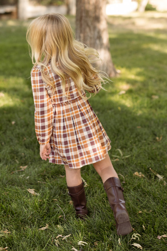 A young girl with long, flowing blonde hair, seen from behind, walks through a grassy field. She is wearing the Ivy City Co Mini Bowie Long Sleeve Dress in Spiced Plaid, featuring shades of brown, orange, and white, paired with knee-high dark brown boots.