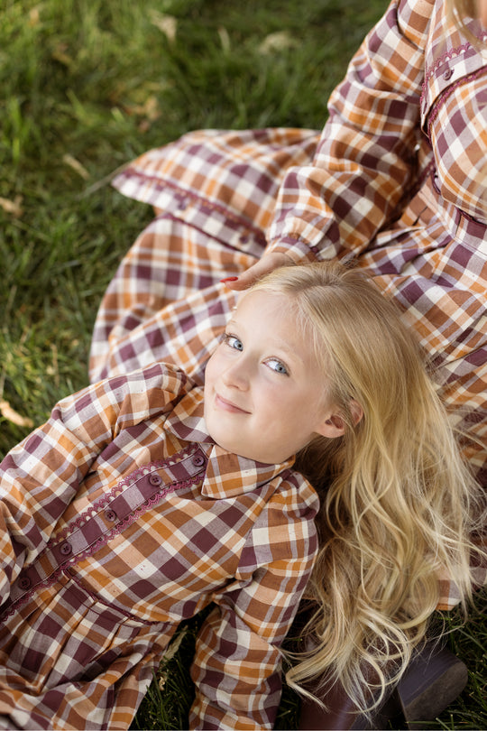 A young girl with long blonde hair wears the Mini Bowie Long Sleeve Dress in Spiced Plaid by Ivy City Co as she lies on the grass, smiling up at the camera. Beside her, another person in a matching Mini Bowie Long Sleeve Dress is partially visible.