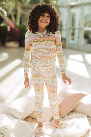 A smiling, curly-haired child stands on a blanket indoors, wearing the Ivy City Co Mini So Soft Pajama Set in the Boxwood Blossoms print. Sunlight pours through large windows behind, with pillows scattered around.