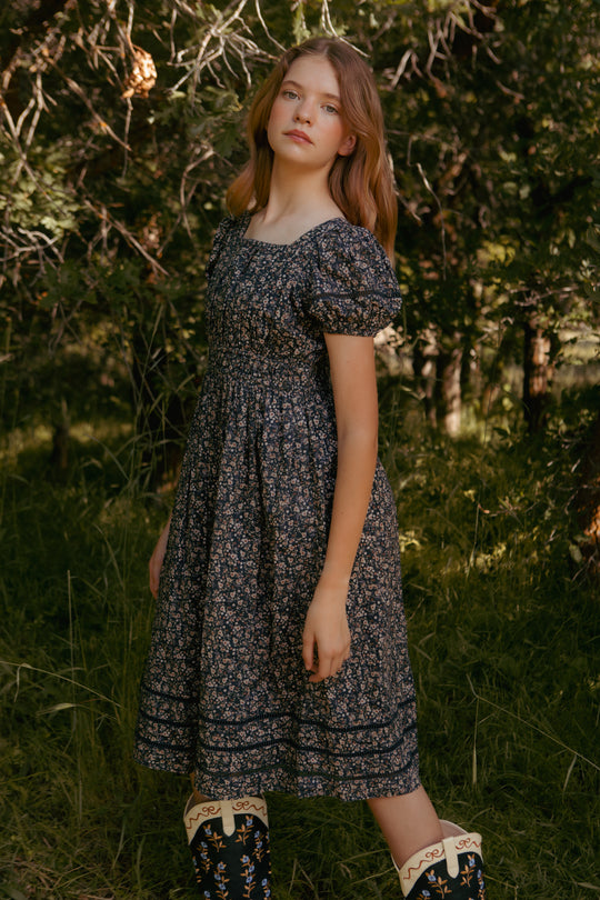 teenager in a floral dress standing in a natural setting with trees and grass.