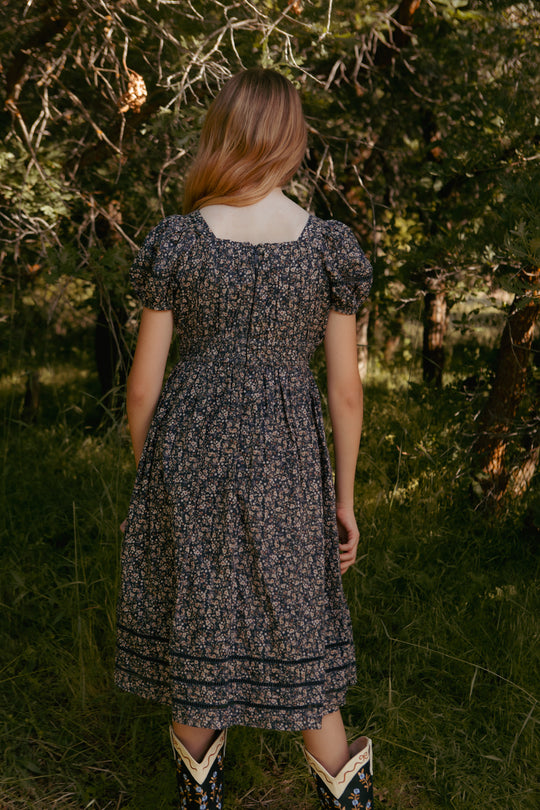 teenager in a floral dress standing in a natural setting with trees and grass.