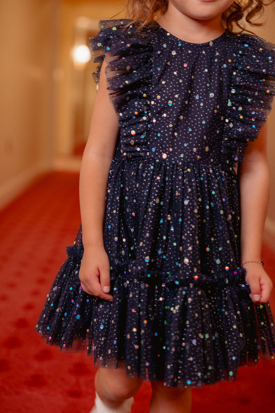 Child wearing a dark blue dress with colorful polka dots on a red carpet.