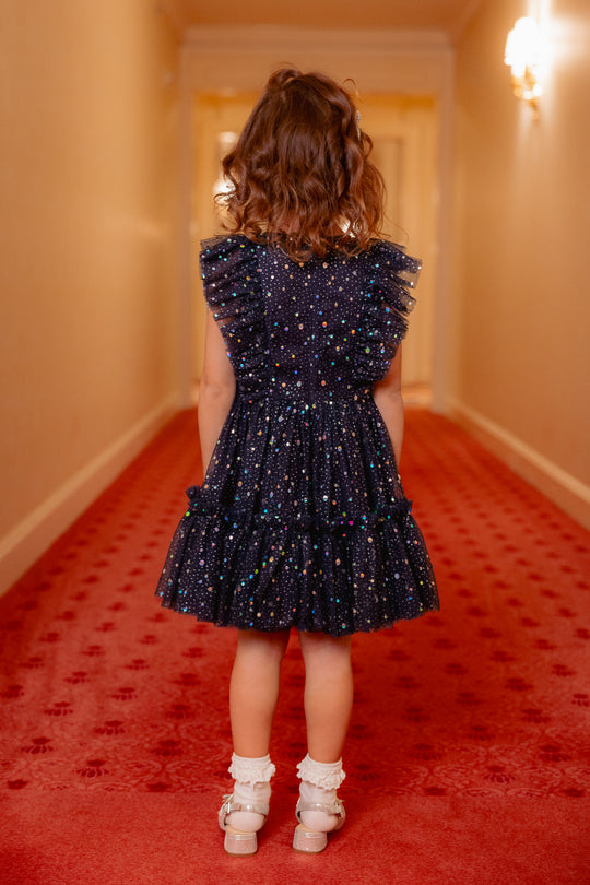 Child in a dark blue dress with white polka dots standing on a red carpeted hallway.