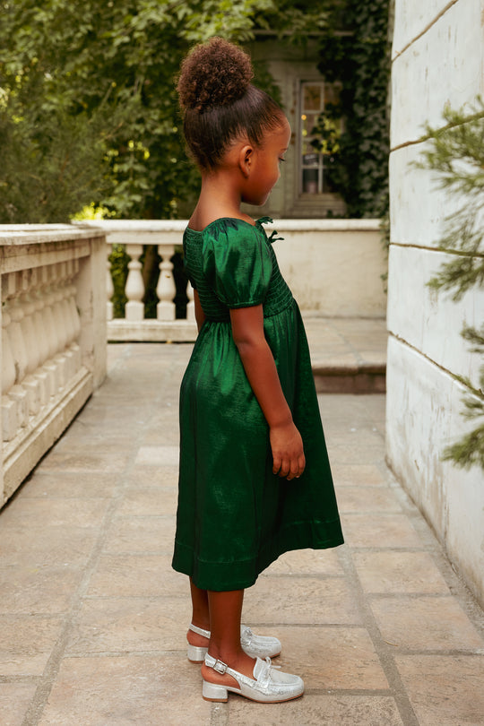 Young girl in a green dress standing on a stone path with trees and a building in the background.