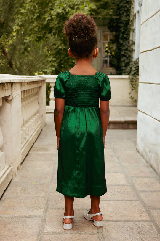 Child in a green dress standing on a stone pathway with trees and a building in the background.