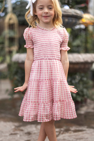 A young blonde girl with a white bow smiles in the Ivy City Co Mini Cordelia Dress in Desert Rose Gingham, featuring puff sleeves and a tiered skirt, while standing outdoors surrounded by greenery.