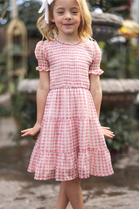 A young blonde girl with a white bow smiles in the Ivy City Co Mini Cordelia Dress in Desert Rose Gingham, featuring puff sleeves and a tiered skirt, while standing outdoors surrounded by greenery.