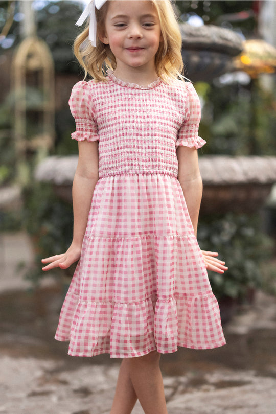 A young blonde girl with a white bow smiles in the Ivy City Co Mini Cordelia Dress in Desert Rose Gingham, featuring puff sleeves and a tiered skirt, while standing outdoors surrounded by greenery.