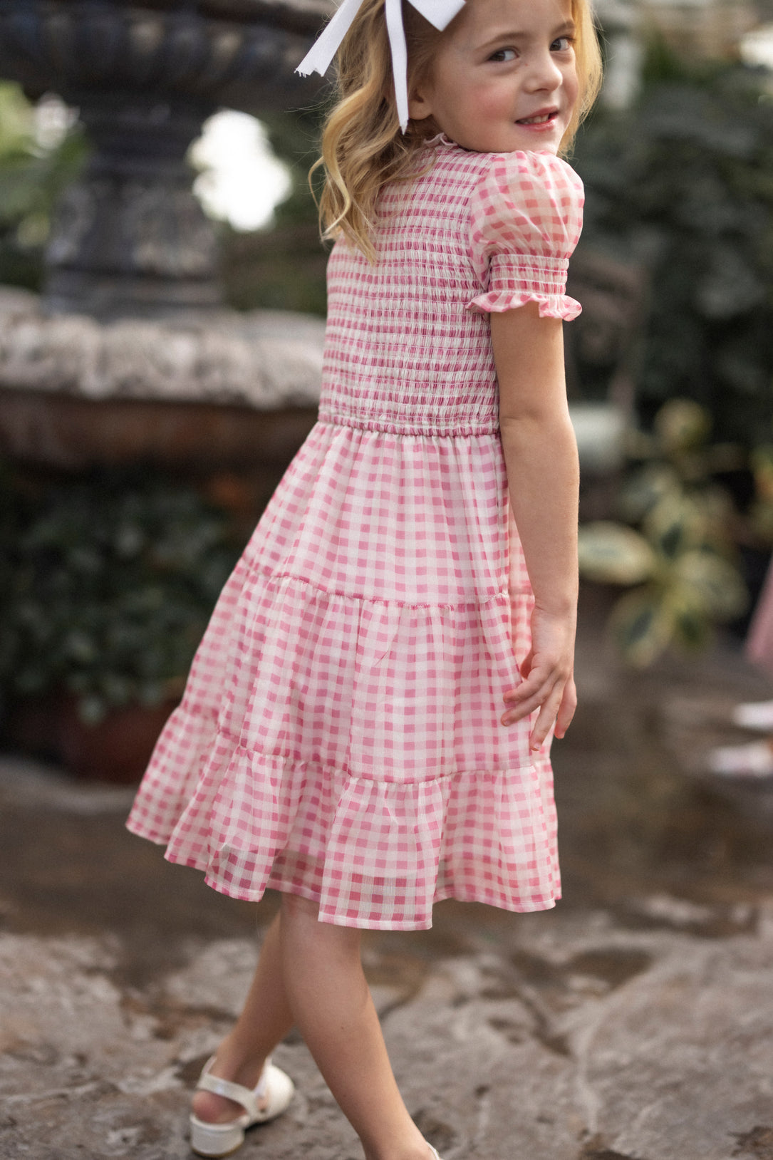 A young girl in the Ivy City Co Mini Cordelia Dress in Desert Rose Gingham, with a white hair bow, smiles over her shoulder while standing outside by a stone fountain.