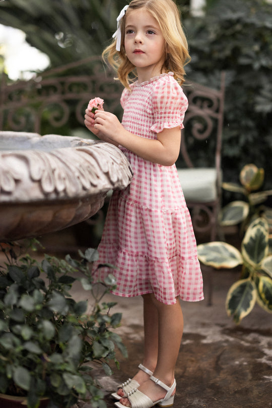 A young girl with light hair and a white bow stands by a stone fountain, holding a pink flower. She wears Ivy City Co's Mini Cordelia Dress in Desert Rose Gingham and white sandals, surrounded by lush green garden plants.