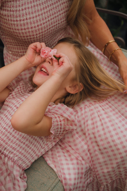 A young girl in the Ivy City Co Mini Cordelia Dress in Desert Rose Gingham playfully holds pink flowers over her eyes while lying on a woman's lap. Both wear matching dresses as the woman gently rests her hand on the girl's side.