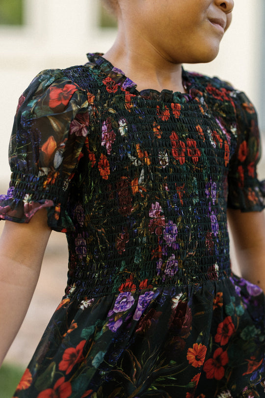 A child wearing the Ivy City Co Mini Cordelia Dress in Evergreen Vivid Meadow—with a smocked bodice and pretty puff sleeves—stands outdoors in a dark floral print.