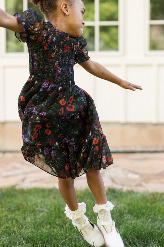 A young girl in the Ivy City Co Mini Cordelia Dress in Evergreen Vivid Meadow, featuring a smocked bodice and puff sleeves, is mid-jump on grass with her arms outstretched and head turned to the side in front of a light-colored building.
