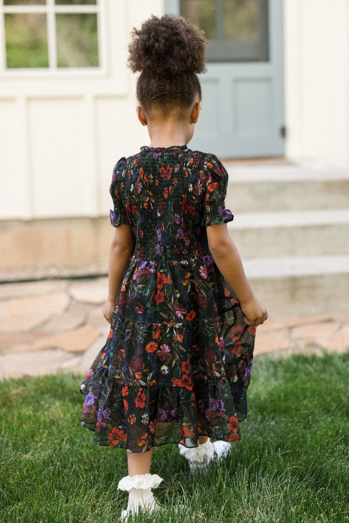 A young child with curly hair stands outside, facing away, wearing the Ivy City Co Mini Cordelia Dress in Evergreen Vivid Meadow. The dress features a smocked bodice and puff sleeves. She pairs it with white socks and holds the skirt of her dress while standing on a grassy lawn in front of stone steps and a light-colored house with a blue door.